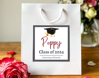 A white luxury graduation gift bag with a black graduation cap design and a custom sticker that reads 'Poppy Class of 2023 University of Lincoln'. The bag is displayed on a wooden surface with flowers in the background.