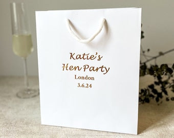 A white hen party gift bag with gold text and detailing, displayed next to a glass of champagne and decorative foliage.