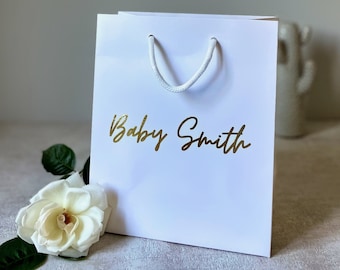 A white luxury gift bag with printing, accompanied by a small white flower and rope handles. Best for New Baby Gift Bag
