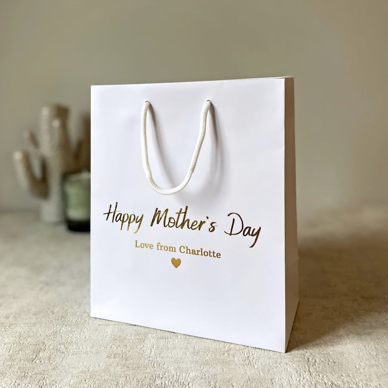 A white gift bag with 'mothers day gift bag' text in gold foil, customizable with a name, presented on a soft surface with a decorative cactus in the background.