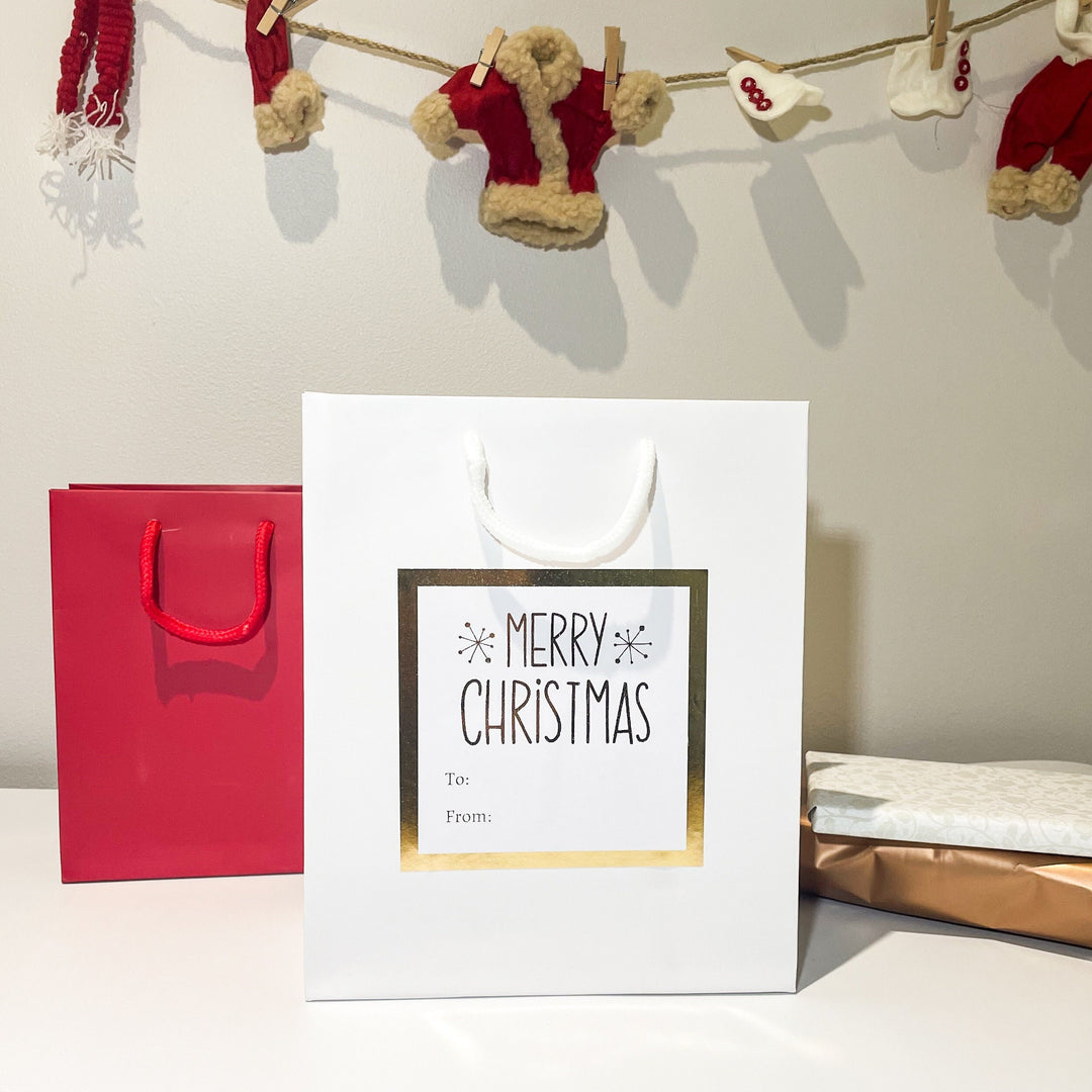 Personalised Christmas gift bags featuring a white bag with a gold-framed 'Merry Christmas' label, perfect for holiday gifting. The bag is accompanied by a red gift bag and wrapped presents, all set against a festive backdrop with Christmas decorations