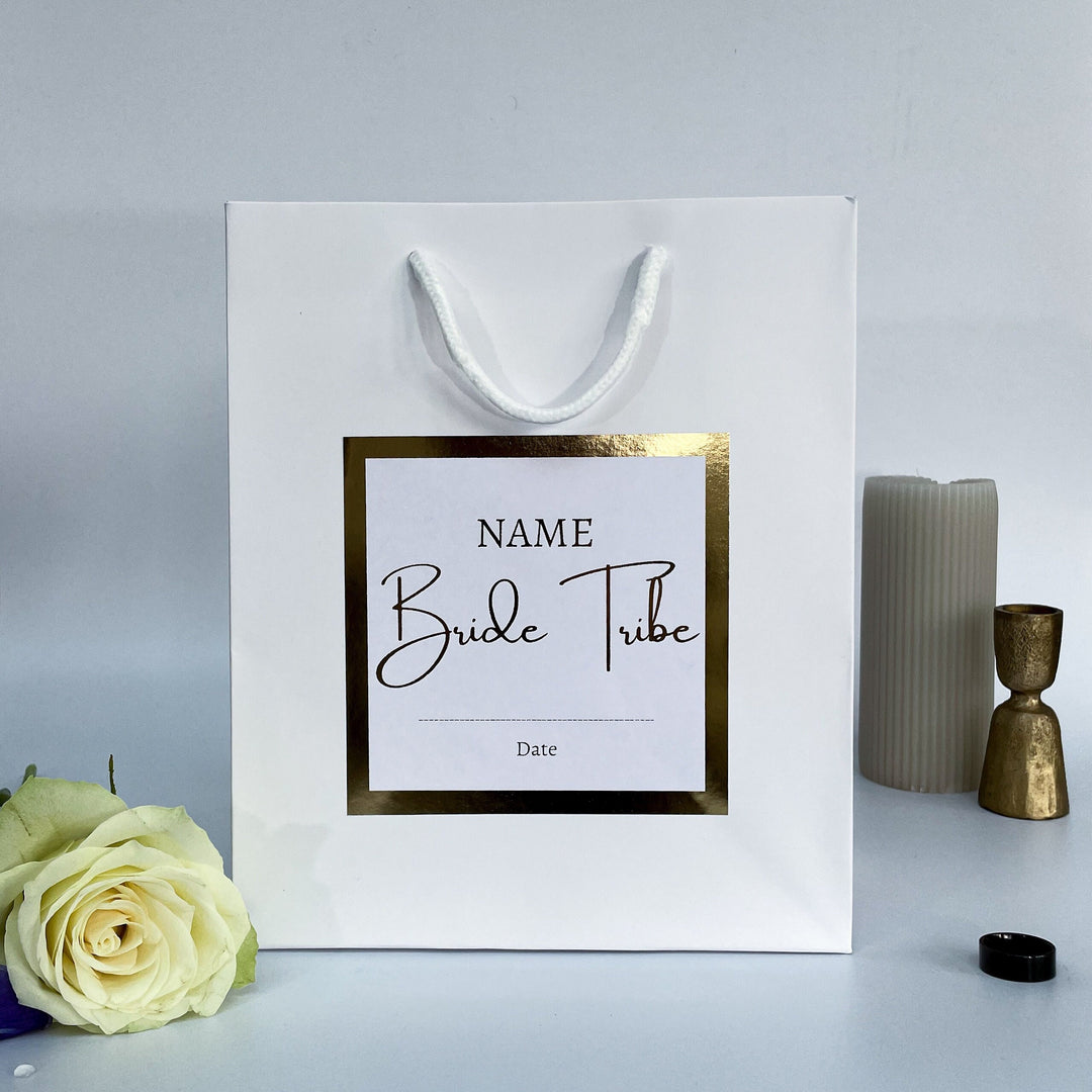 A white personalized gift bag for a Hen Party, featuring a gold-framed label with customizable fields for "NAME" and "Date." The text "Bride Tribe" is prominently displayed in a stylish font. The bag is surrounded by a rose and elegant decorative items.