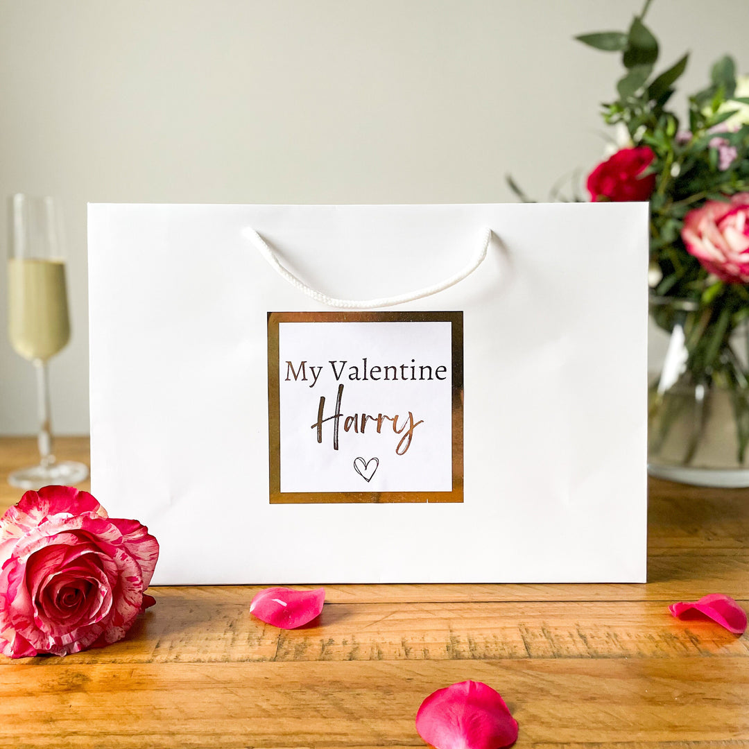 "Valentine's gift bag with the text 'My Valentine Harry' on a white bag, surrounded by rose petals, a glass of champagne, and a vase of flowers in the background."