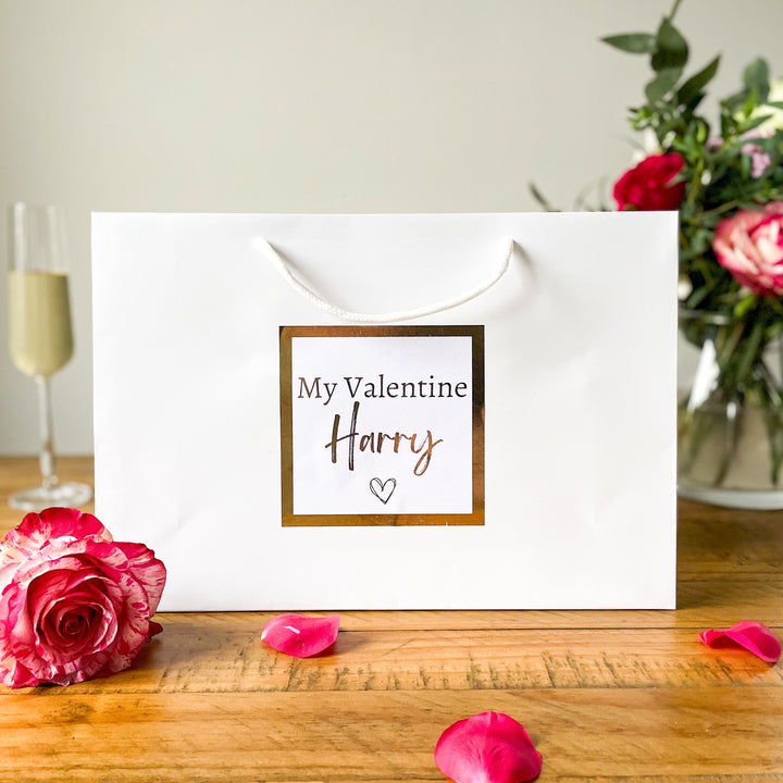 "Valentine's gift bag with the text 'My Valentine Harry' on a white bag, surrounded by rose petals, a glass of champagne, and a vase of flowers in the background."