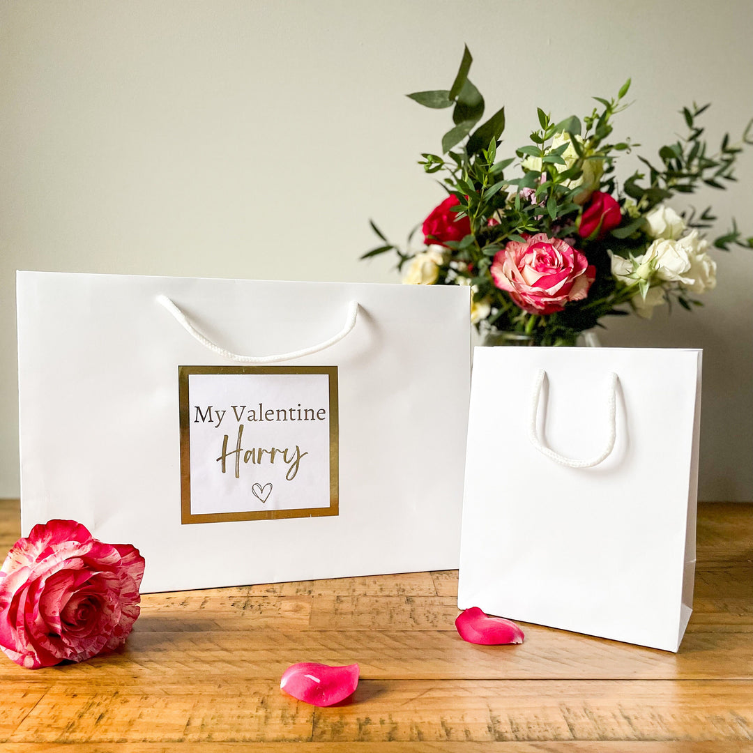 "Valentine's gift bag with the text 'My Valentine Harry' on a white bag, surrounded by rose petals, a glass of champagne, and a vase of flowers in the background."