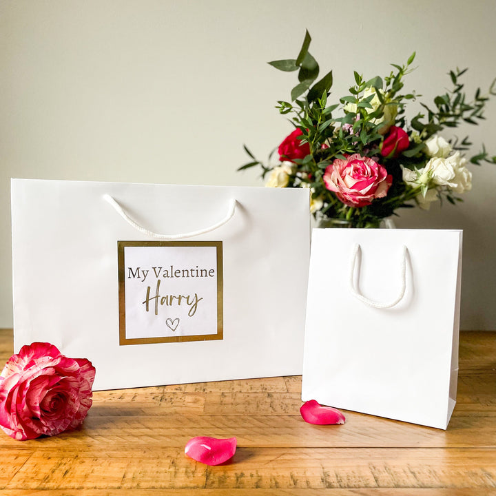 "Valentine's gift bag with the text 'My Valentine Harry' on a white bag, surrounded by rose petals, a glass of champagne, and a vase of flowers in the background."