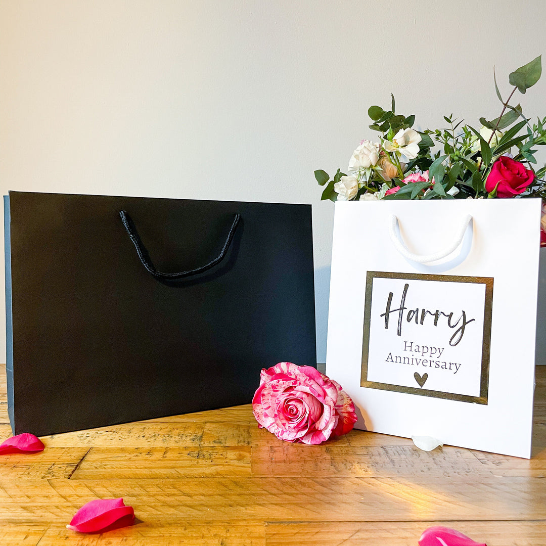 A large balck and a white Anniversary gift bag with a 'Happy Anniversary' label featuring the name 'Harry' in elegant, gold script. The label is bordered with a gold square frame and a small heart at the bottom. The bag is placed on a wooden table, with a pink rose in the foreground and a soft floral arrangement visible in the background.