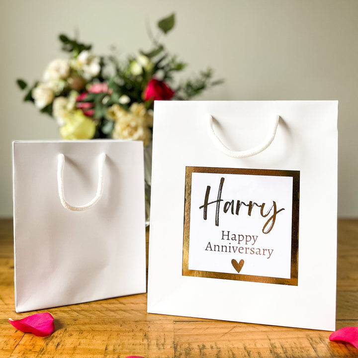 A white Anniversary gift bag with a 'Happy Anniversary' label featuring the name 'Harry' in elegant, gold script. The label is bordered with a gold square frame and a small heart at the bottom. The bag is placed on a wooden table, with a pink rose in the foreground and a soft floral arrangement visible in the background.