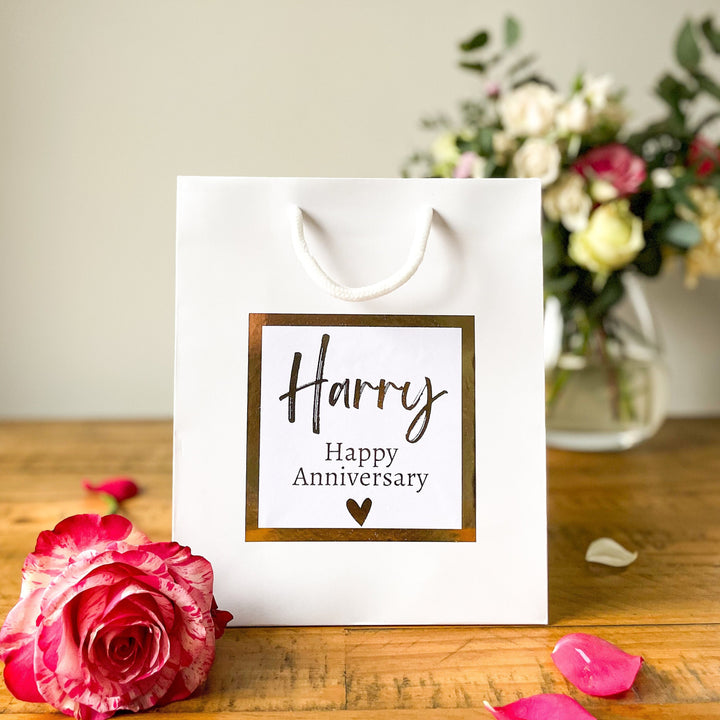 A white Anniversary gift bag with a 'Happy Anniversary' label featuring the name 'Harry' in elegant, gold script. The label is bordered with a gold square frame and a small heart at the bottom. The bag is placed on a wooden table, with a pink rose in the foreground and a soft floral arrangement visible in the background.