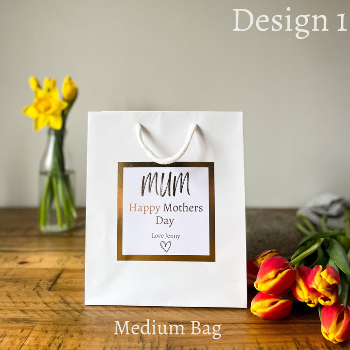 White gift bag with gold-bordered “Happy Mother’s Day” sticker, personalised with “Love Jenny”, placed on a wooden table with yellow daffodils in a vase and red-yellow tulips nearby.
