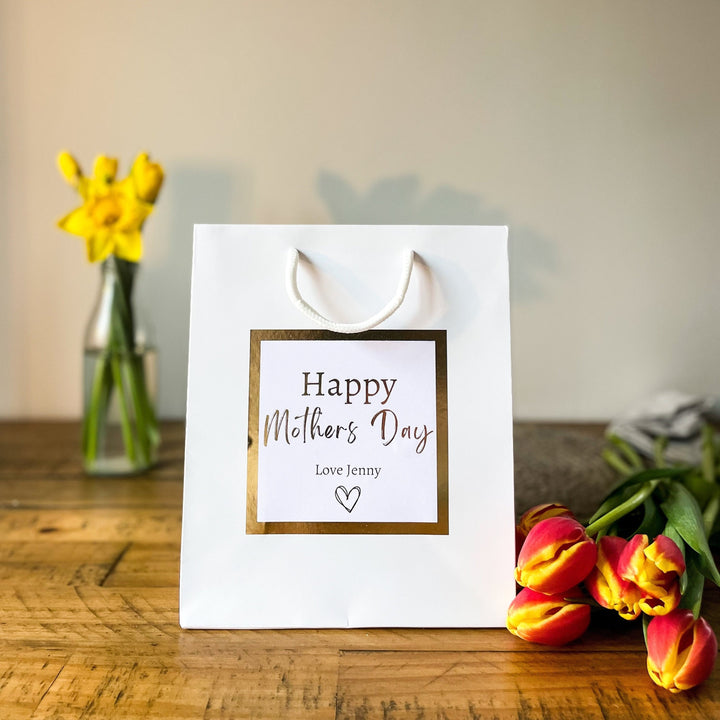 White mother's day gift bag with gold-bordered “Happy Mother’s Day” sticker, personalised with “Love Jenny”, placed on a wooden table with yellow daffodils in a vase and red-yellow tulips nearby.