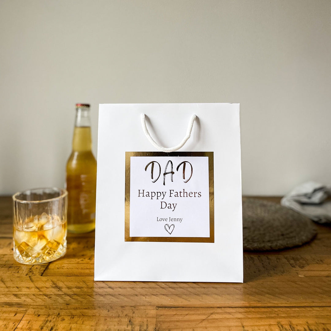 A white father's day gift bags with a gold-trimmed label that reads "DAD Happy Father's Day" and is signed "Love Jenny."