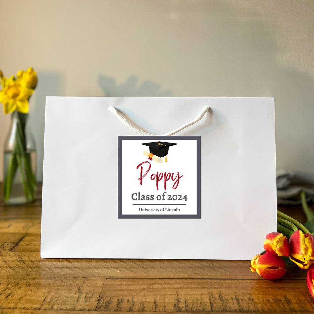 A large white luxury graduation gift bag with a black graduation cap design and a custom sticker that reads 'Poppy Class of 2023 University of Lincoln'. The bag is displayed on a wooden surface with flowers in the background.