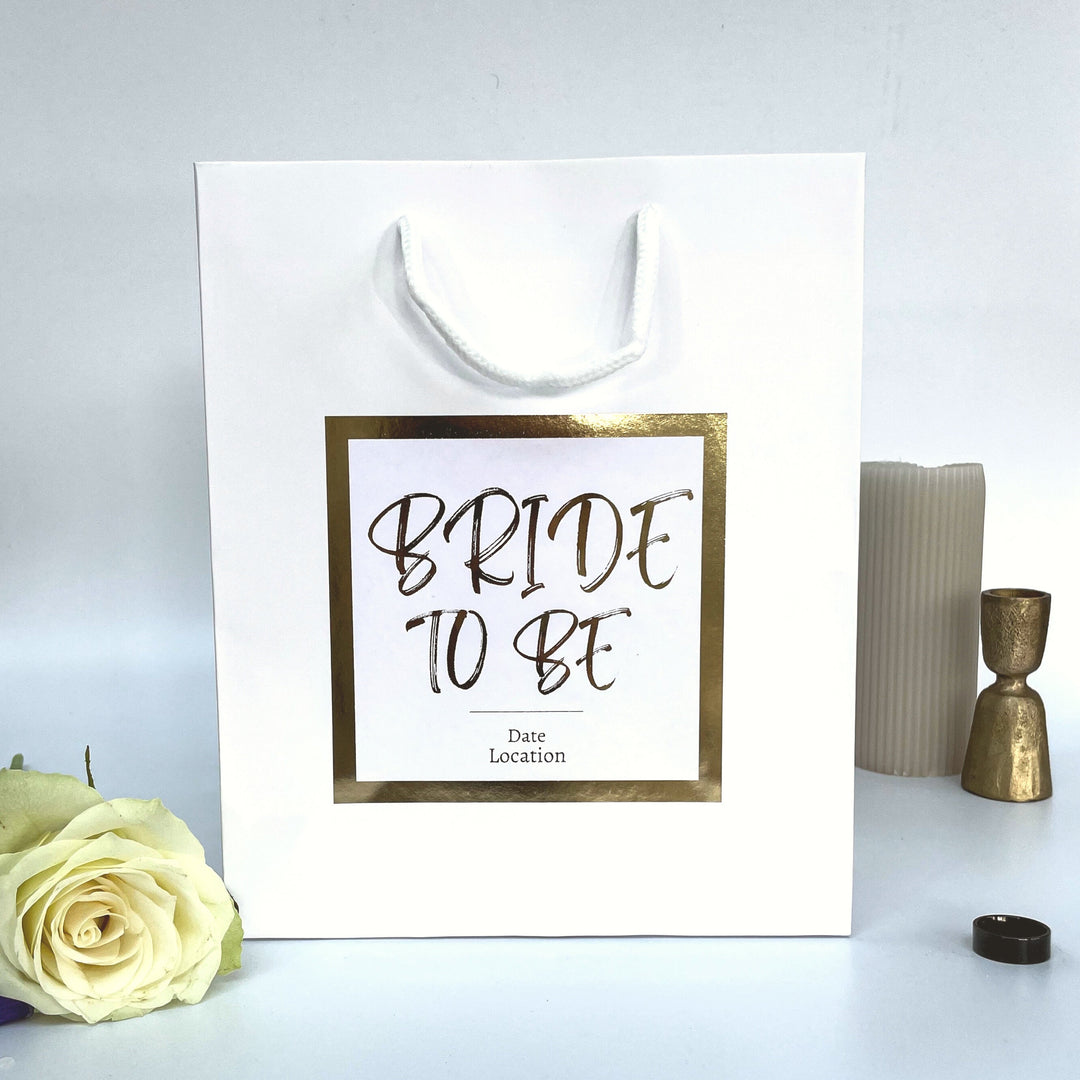 A "Bride to Be" gift bag with a gold foil border around the label. The label features the words "BRIDE TO BE" in bold, elegant font, with the text "Date" and "Location" underneath. The bag is white, placed next to a white rose, a candle, and a gold candlestick.