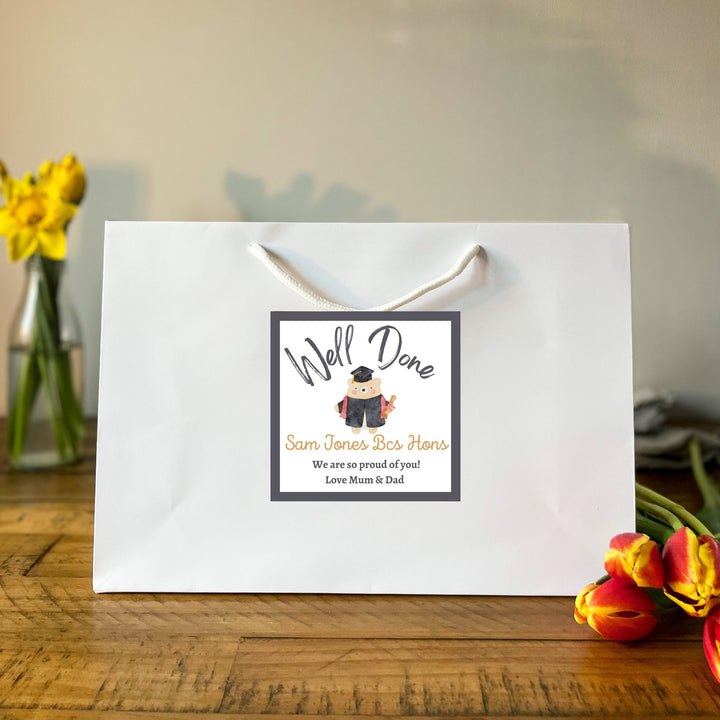A white graduation gift bag with a 'Well Done' sticker featuring text and an illustration of a graduation cap. The sticker is designed for personalisation with names and messages.