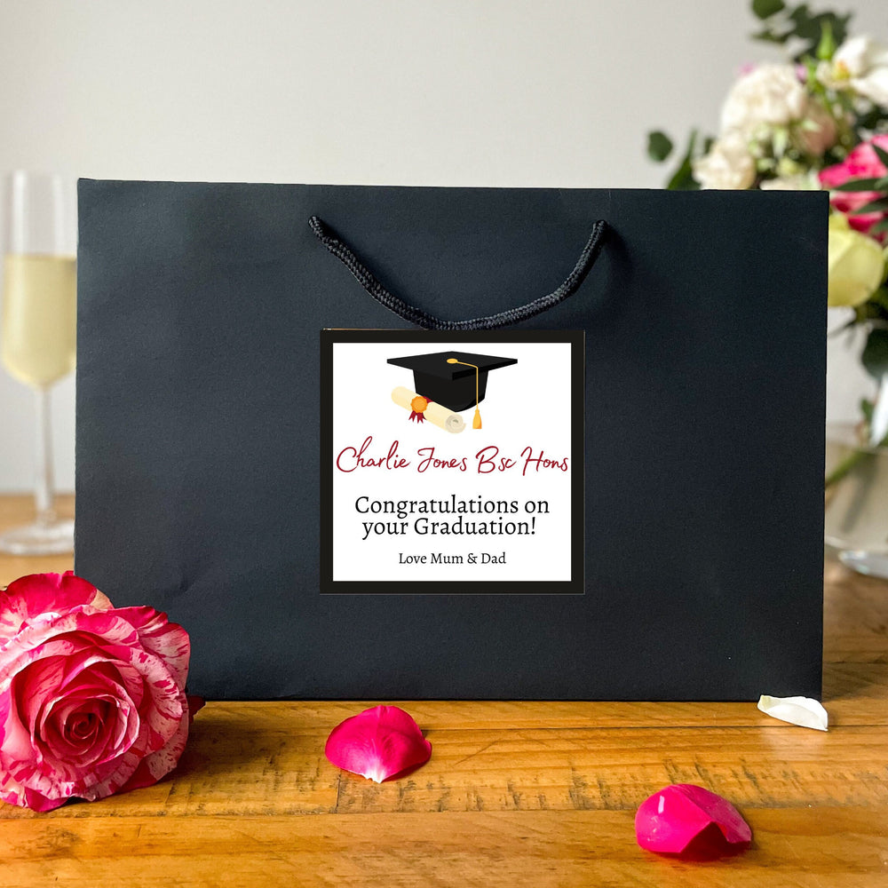 A Black Graduation Gift Bag with a square label featuring the message "Charlie Jones Be Honours" in red and black text, along with a graduation cap graphic. Below the name, it reads "Congratulations on your Graduation!" followed by "Love Mum & Dad." The bag is placed on a wooden table, surrounded by vibrant yellow daffodils and red tulips in the background, creating a celebratory and warm atmosphere.