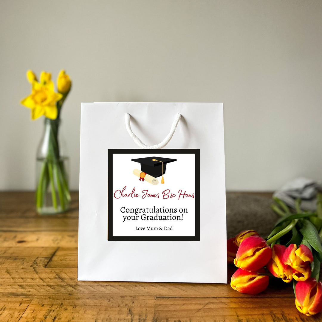 A white Graduation Gift Bag with a square label featuring the message "Charlie Jones Be Honours" in red and black text, along with a graduation cap graphic. Below the name, it reads "Congratulations on your Graduation!" followed by "Love Mum & Dad." The bag is placed on a wooden table, surrounded by vibrant yellow daffodils and red tulips in the background, creating a celebratory and warm atmosphere.