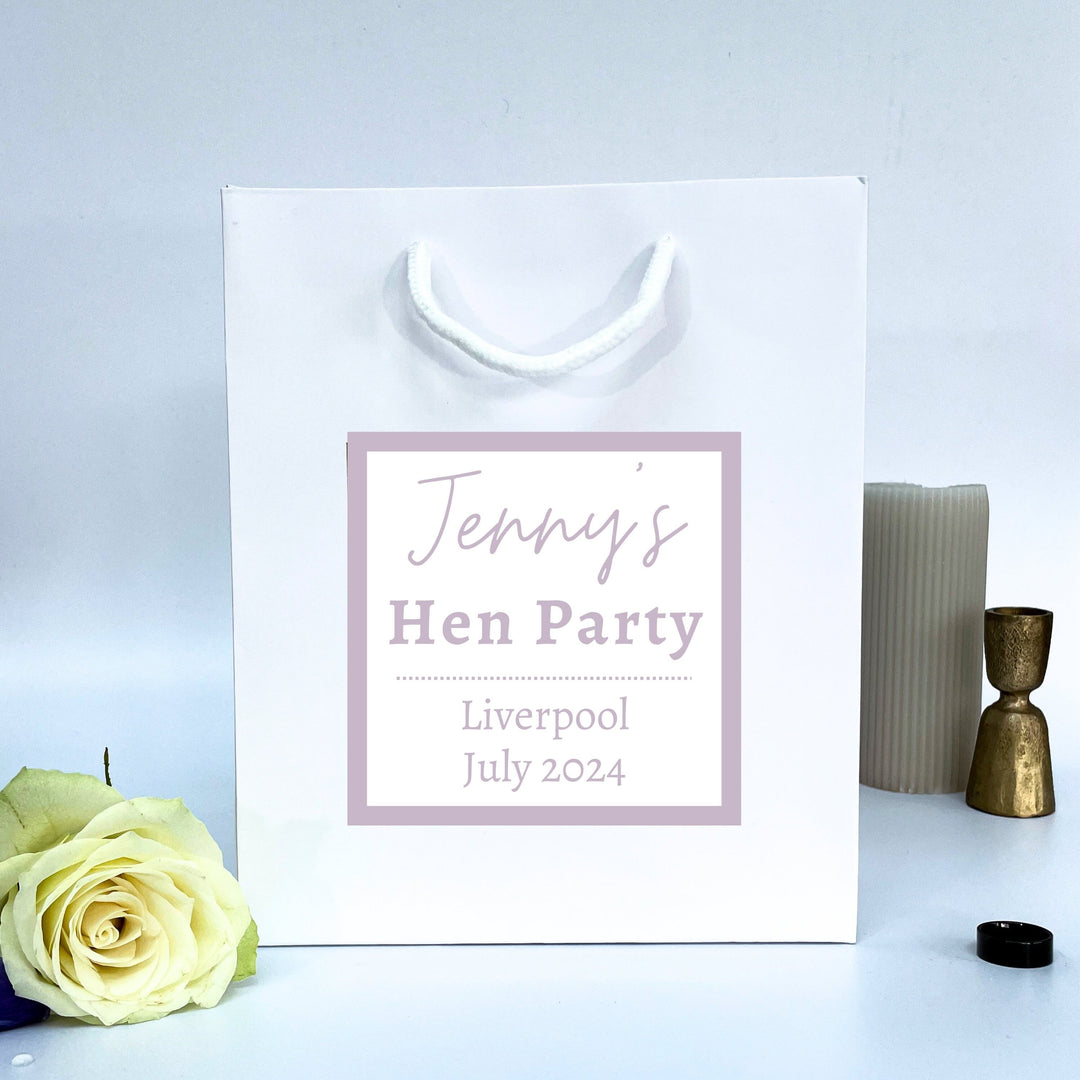 A white Hen Party Gift Bags with a label that reads "Jenny's Hen Party" in a light purple font, along with the text "Liverpool July 2024" below it. The bag is placed on a surface next to a white rose, a candle, and a small black object.