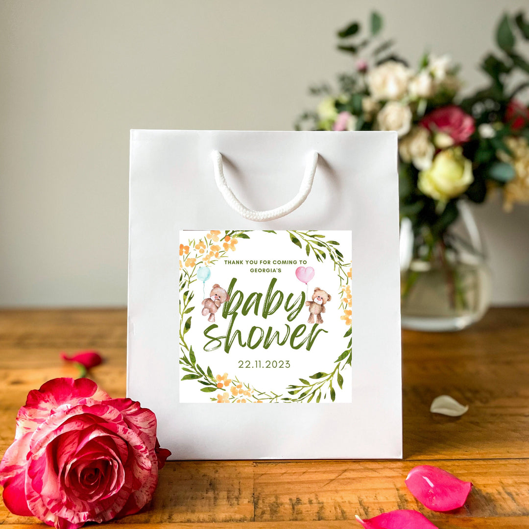 A white gift bag with a colorful 'Thank you for coming to Georgia's baby shower gift bags' label, featuring a design with teddy bears, balloons, and floral elements. The date '22.11.2023' is also displayed. The bag is placed on a wooden table with a pink rose nearby, and a soft floral arrangement can be seen in the background.