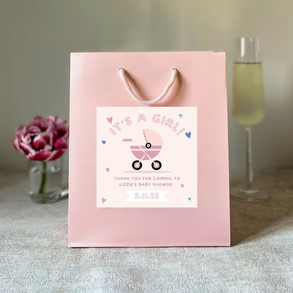 A Pink baby shower party bags gift with 'It's a Boy' sticker, blue handles, and a blue baby shower design, displayed next to a candle and a glass of champagne.