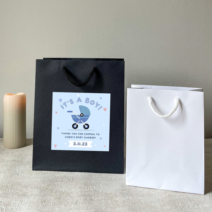 Black and white baby shower party bags gift with 'It's a Boy' sticker, blue handles, and a blue baby shower design, displayed next to a candle and a glass of champagne.