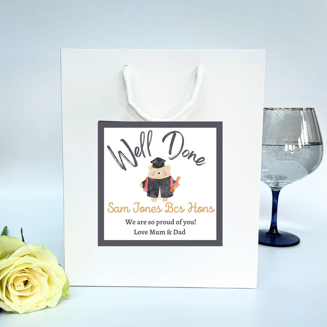 A white graduation gift bag with a 'Well Done' sticker featuring text and an illustration of a graduation cap. The sticker is designed for personalisation with names and messages.