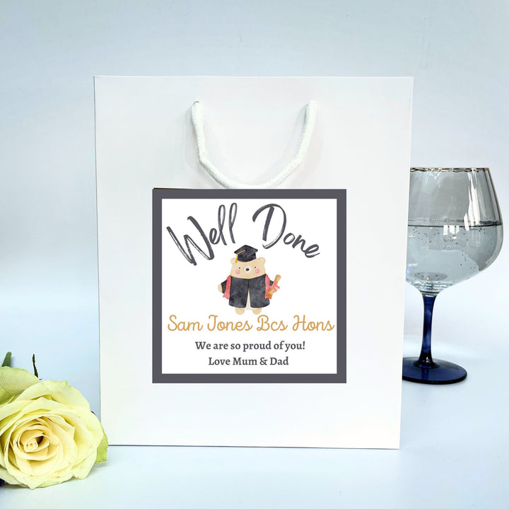 A white graduation gift bag with a 'Well Done' sticker featuring text and an illustration of a graduation cap. The sticker is designed for personalisation with names and messages.