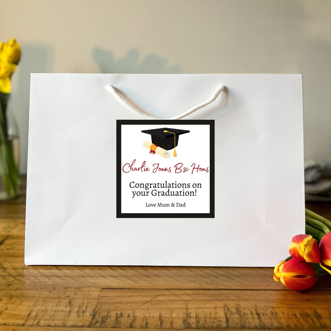 A large white Graduation Gift Bag with a square label featuring the message "Charlie Jones Be Honours" in red and black text, along with a graduation cap graphic. Below the name, it reads "Congratulations on your Graduation!" followed by "Love Mum & Dad." The bag is placed on a wooden table, surrounded by vibrant yellow daffodils and red tulips in the background, creating a celebratory and warm atmosphere.
