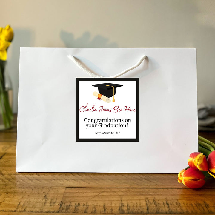 A large white Graduation Gift Bag with a square label featuring the message "Charlie Jones Be Honours" in red and black text, along with a graduation cap graphic. Below the name, it reads "Congratulations on your Graduation!" followed by "Love Mum & Dad." The bag is placed on a wooden table, surrounded by vibrant yellow daffodils and red tulips in the background, creating a celebratory and warm atmosphere.