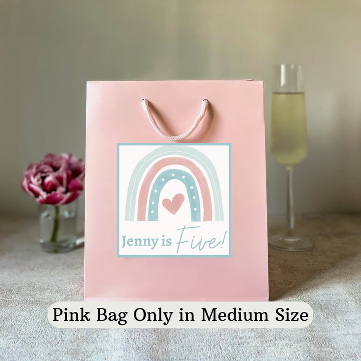 A personalised pink gift rainbow party bag with a rainbow pattern and custom text 'Jenny is Five!' printed on the front, displayed on a table with a decorative cactus in the background