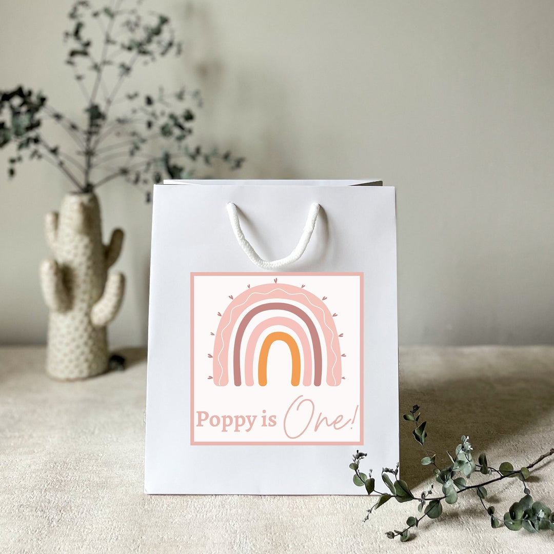 A personalised white gift rainbow party bag with a rainbow pattern and custom text 'Poppy is One!' printed on the front, displayed on a table with a decorative cactus in the background.