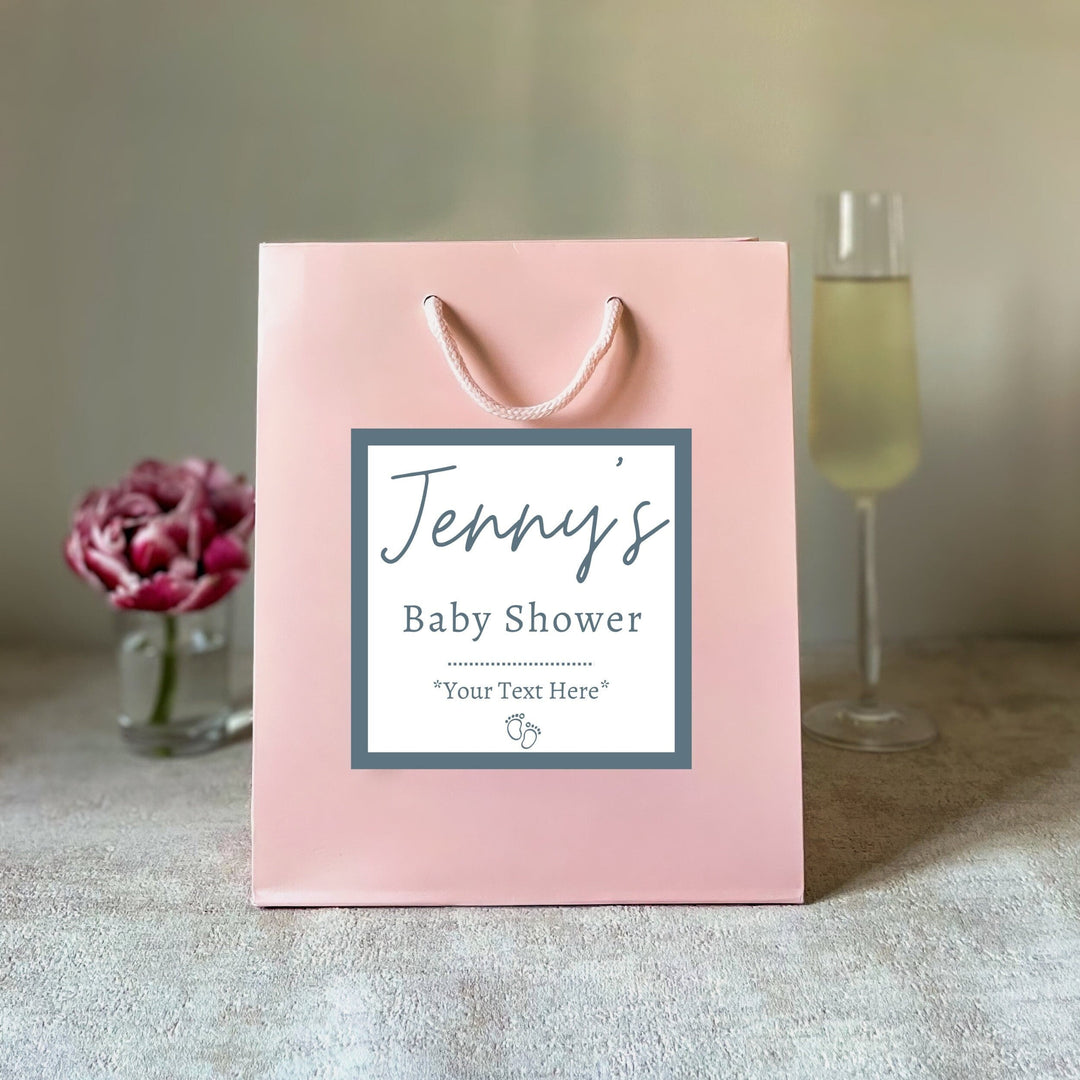 A light pink luxury baby shower gift bag uk with a matte finish, featuring a personalized sticker with the text 'Jenny's Baby Shower' placed on a table with a glass of champagne and a flower in the background.