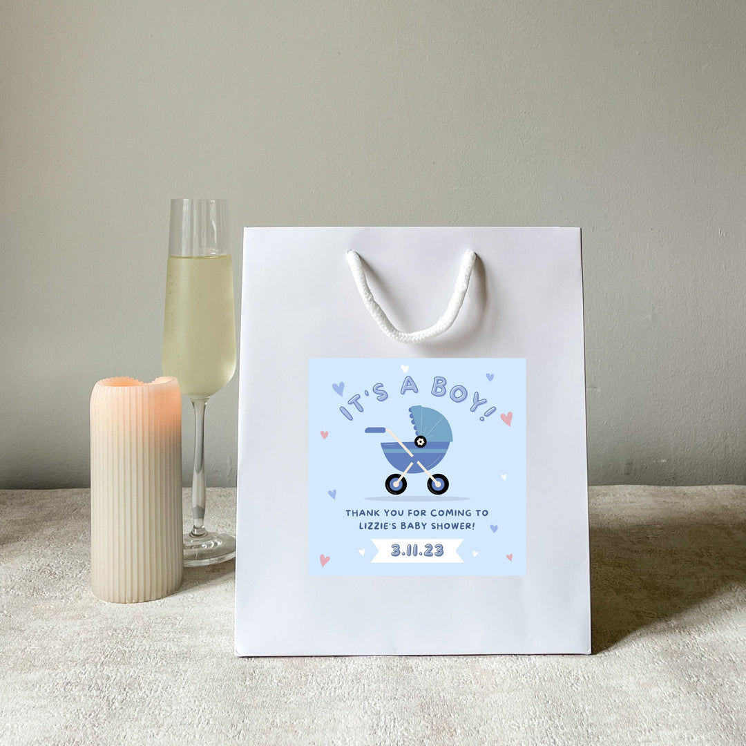 A white baby shower party bags gift with 'It's a Boy' sticker, blue handles, and a blue baby shower design, displayed next to a candle and a glass of champagne.