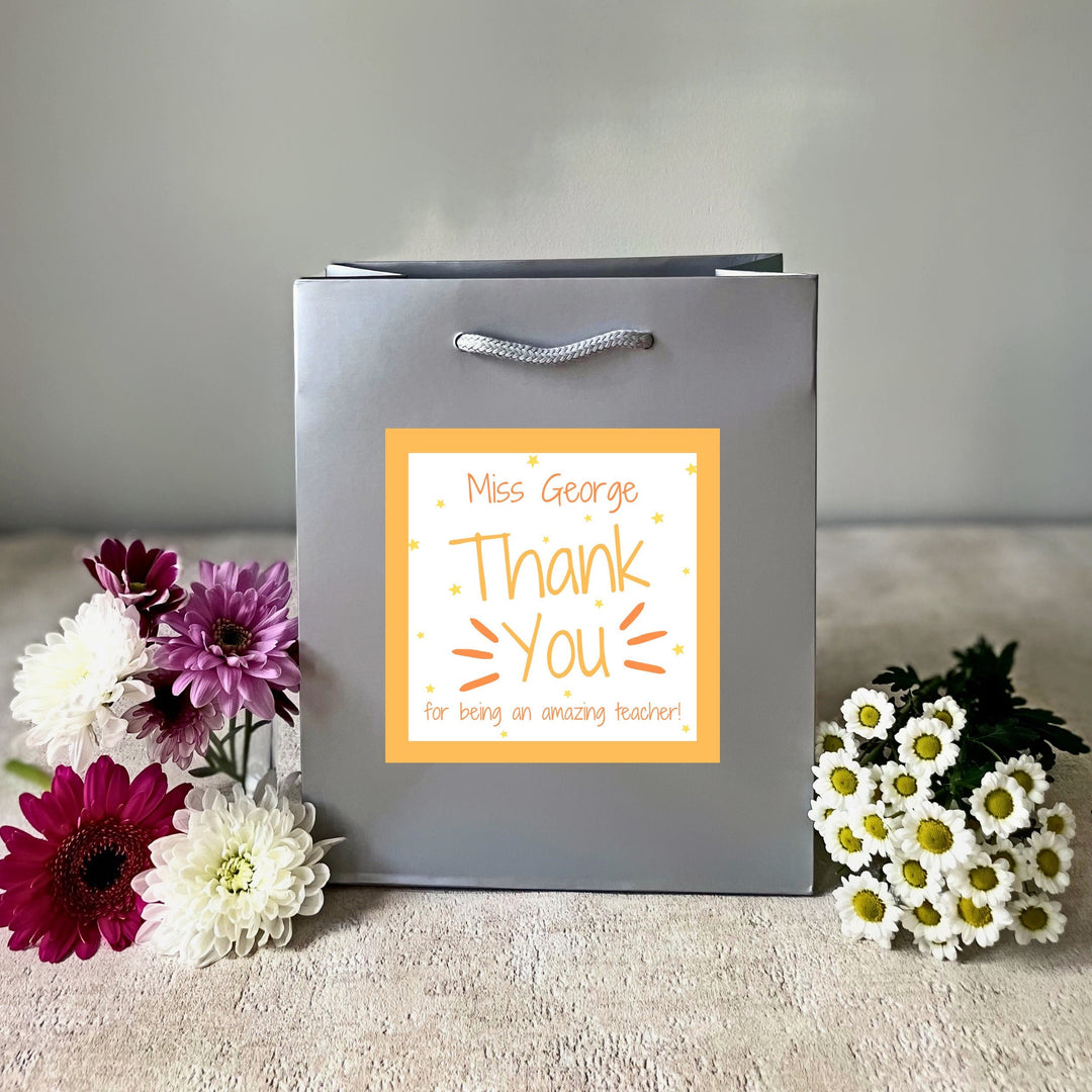 A silver luxury teacher gift bag with a personalised 'Thank You' sticker attached, presented alongside colourful flowers.