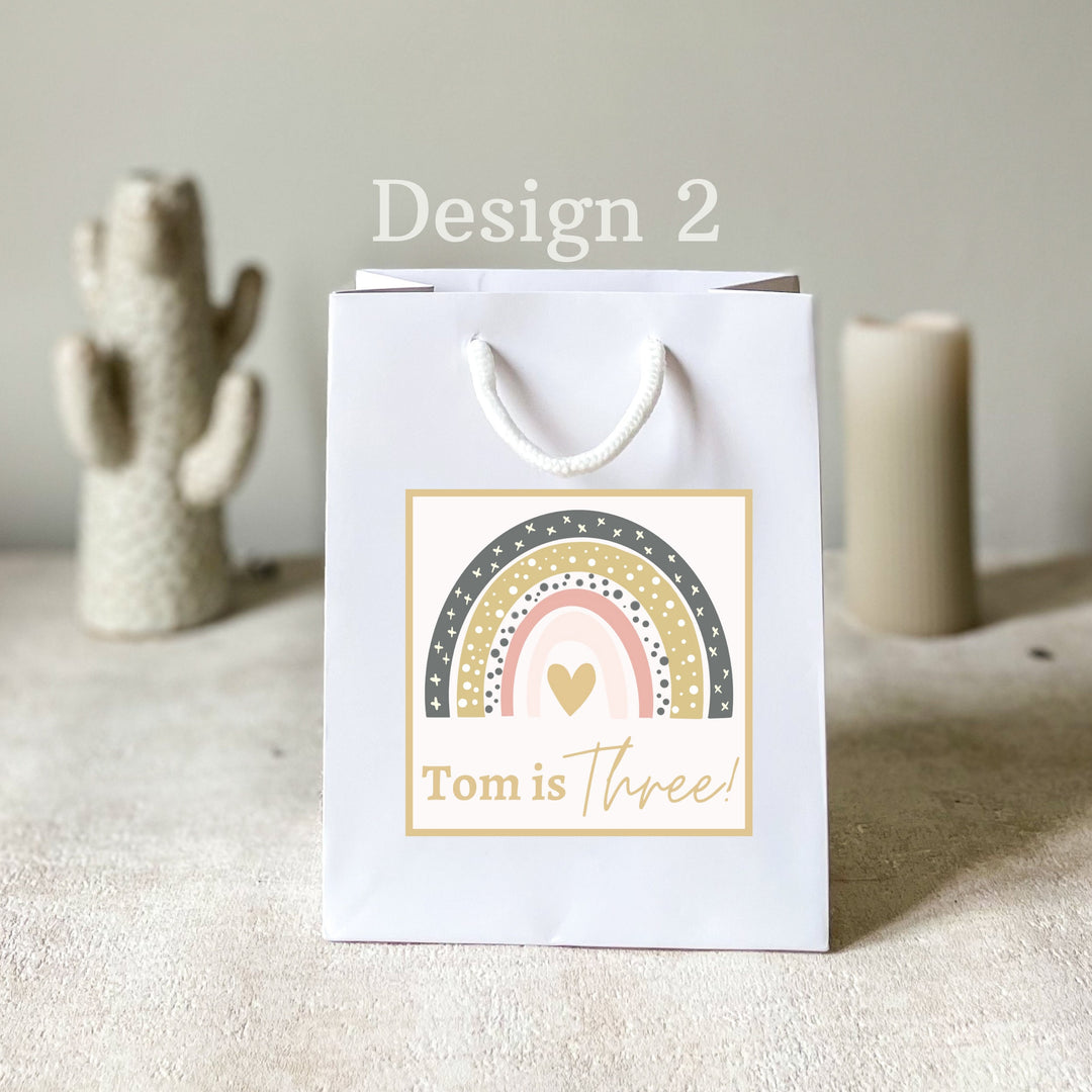 A personalised white gift rainbow party bag with a rainbow pattern and custom text 'tom is three!' printed on the front, displayed on a table with a decorative cactus in the background.