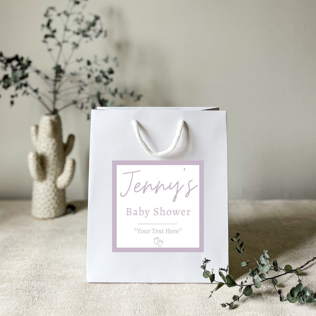 A light matt white luxury baby shower gift bag uk with a matte finish, featuring a personalized sticker with the text 'Jenny's Baby Shower' placed on a table with a glass of champagne and a flower in the background.