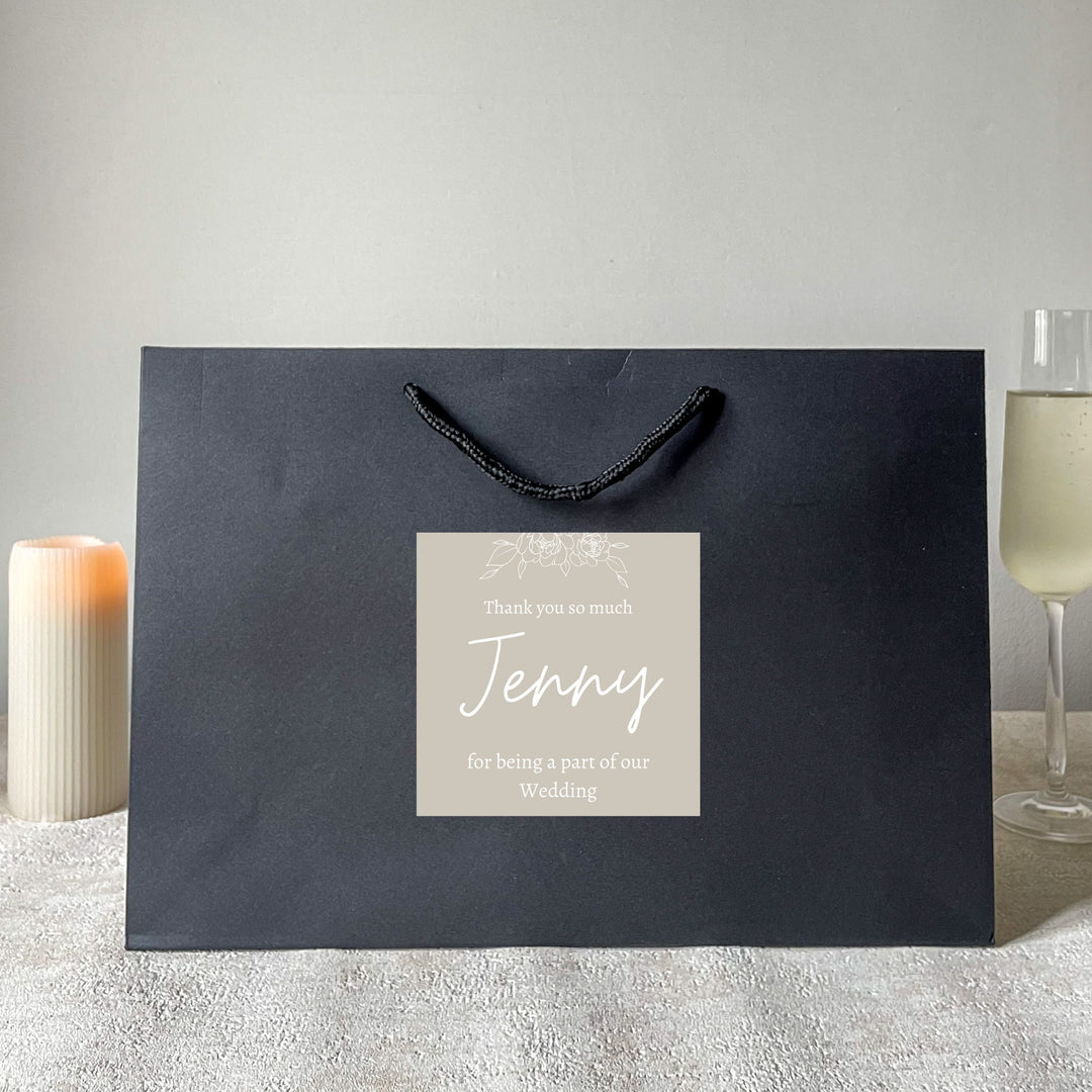 A large black bridesmaids gift bags with a matte finish, featuring a custom sticker that thanks Jenny for being a part of the wedding. The sticker is green with white text, and the bag is presented on a surface with flowers and a glass of champagne in the background.