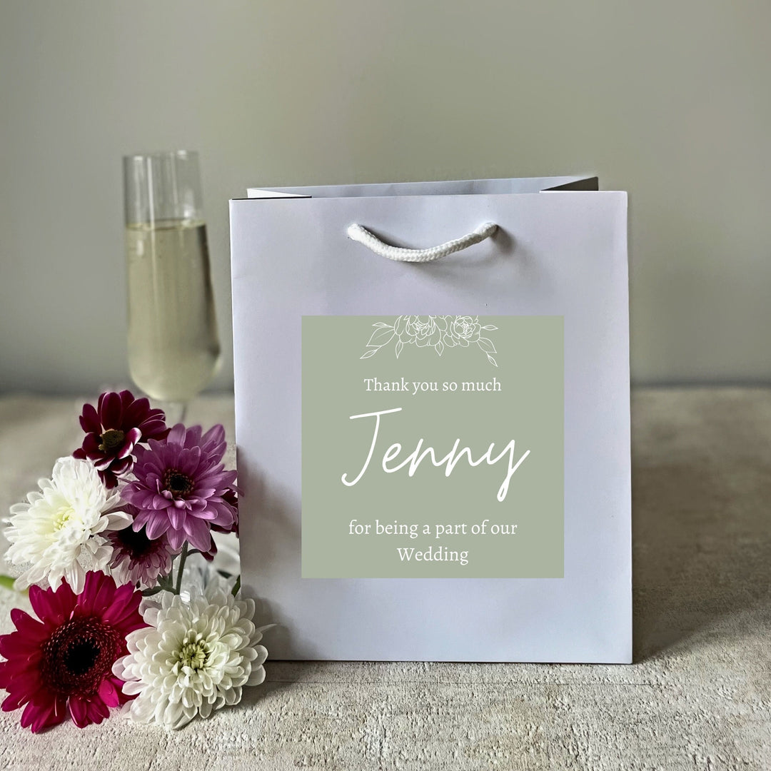 A white bridesmaids gift bags with a matte finish, featuring a custom sticker that thanks Jenny for being a part of the wedding. The sticker is green with white text, and the bag is presented on a surface with flowers and a glass of champagne in the background.