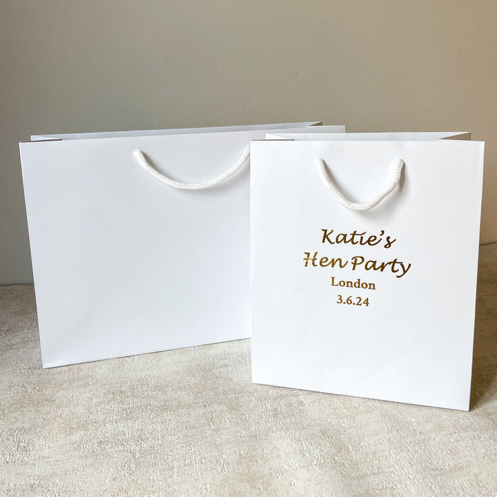 Two white hen party gift bag with gold text and detailing, displayed next to a glass of champagne and decorative foliage.