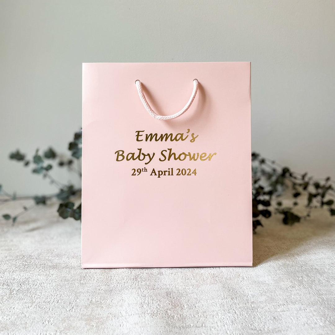 A pink gift bag with 'Emma's Baby Shower' printed in gold foil, with a date '29 april 2024' below the name. The bag is closed with a string and has a handle.