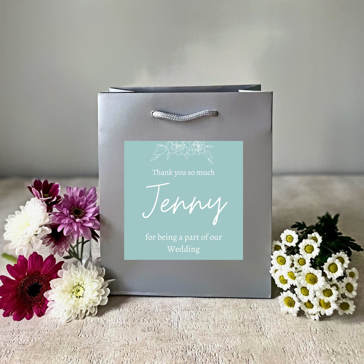 A grey bridesmaids gift bags with a matte finish, featuring a custom sticker that thanks Jenny for being a part of the wedding. The sticker is green with white text, and the bag is presented on a surface with flowers and a glass of champagne in the background.