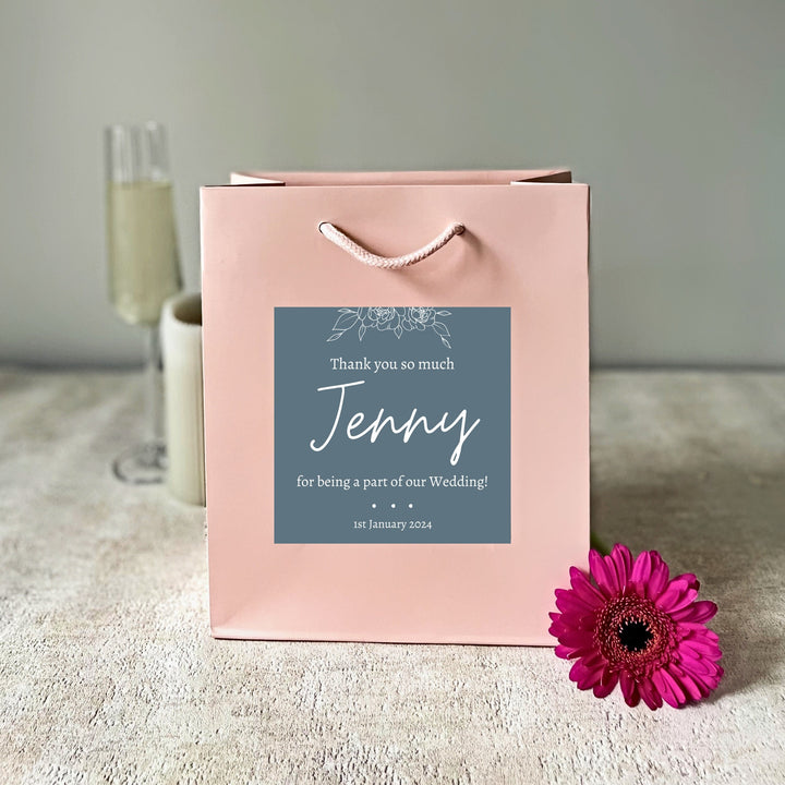 A pink bridesmaids gift bags with a matte finish, featuring a custom sticker that thanks Jenny for being a part of the wedding. The sticker is green with white text, and the bag is presented on a surface with flowers and a glass of champagne in the background.
