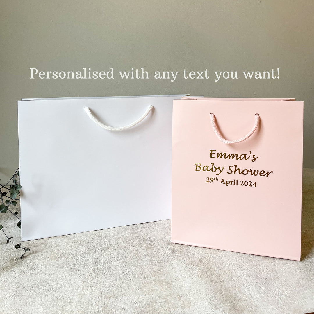 white and pink hen party gift bag with gold text and detailing, displayed next to a glass of champagne and decorative foliage.