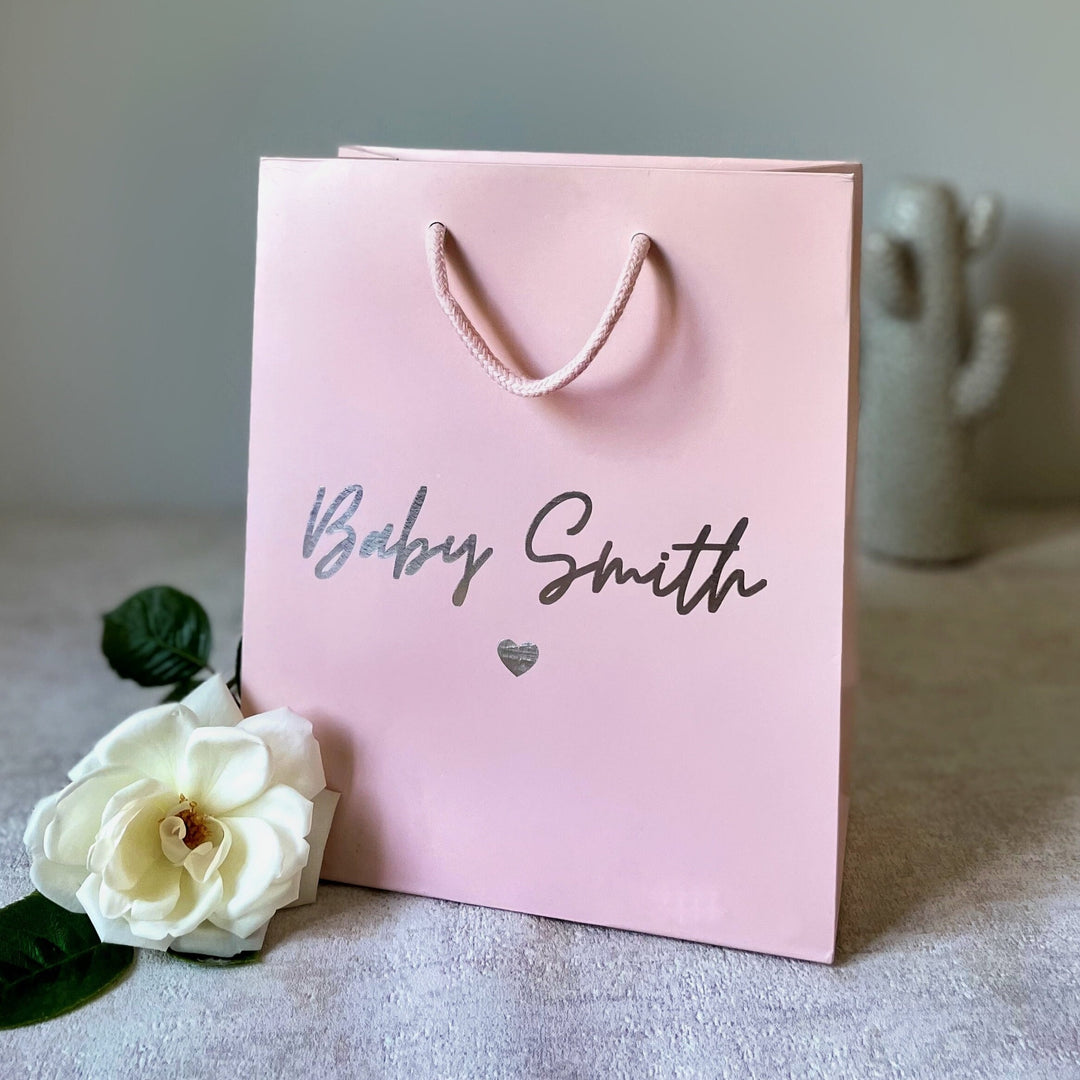 A pink luxury gift bag with printing, accompanied by a small white flower and rope handles. Best for New Baby Gift Bag