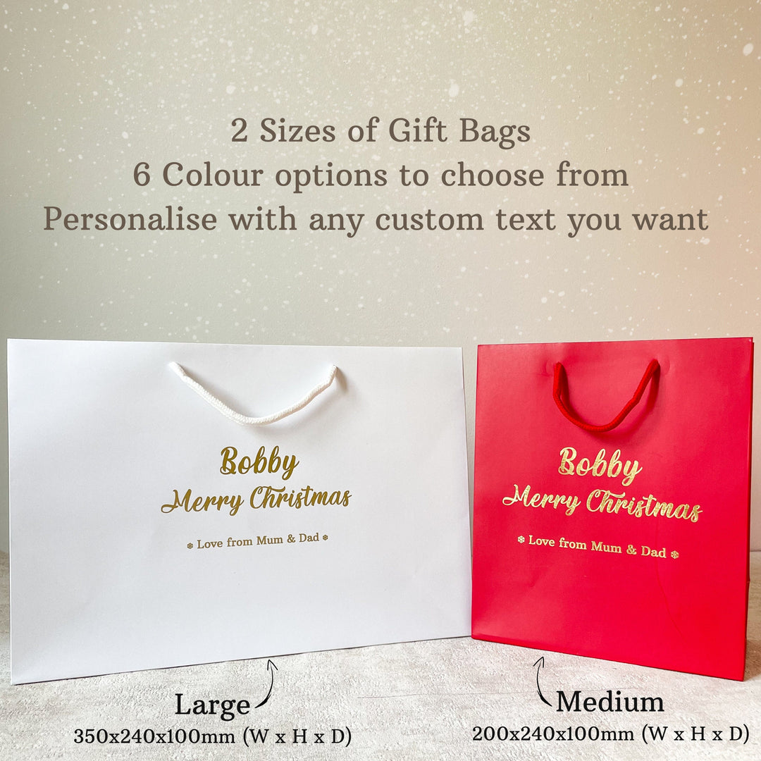 white and red Christmas gift bag with gold foil lettering that reads "Bobby" and "Merry Christmas" on the front. Below the festive message, it says "Love from Mum & Dad" in smaller gold text. The bag features white rope handles and is placed on a neutral-colored surface, with a vase of white flowers in the background, giving it a warm holiday feel.