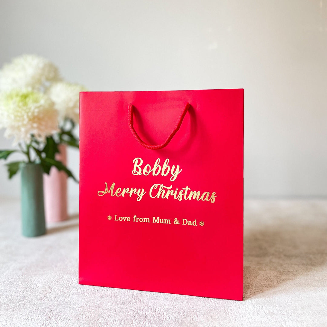 A red luxury rope-handled Christmas gift bag with 'Merry Christmas' printed in gold, placed on a white surface with a decorative element in the background.