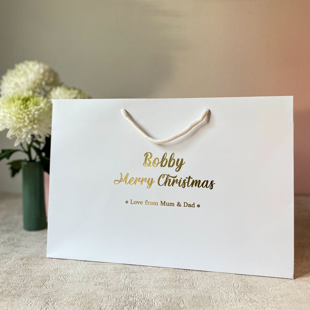 A white Christmas gift bag with gold foil lettering that reads "Bobby" and "Merry Christmas" on the front. Below the festive message, it says "Love from Mum & Dad" in smaller gold text. The bag features white rope handles and is placed on a neutral-colored surface, with a vase of white flowers in the background, giving it a warm holiday feel.