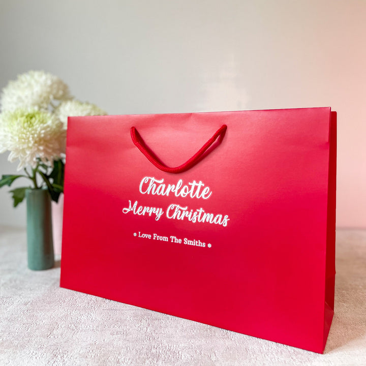 A red Christmas gift bag with gold foil lettering that reads "Bobby" and "Merry Christmas" on the front. Below the festive message, it says "Love from Mum & Dad" in smaller gold text. The bag features white rope handles and is placed on a neutral-colored surface, with a vase of white flowers in the background, giving it a warm holiday feel.