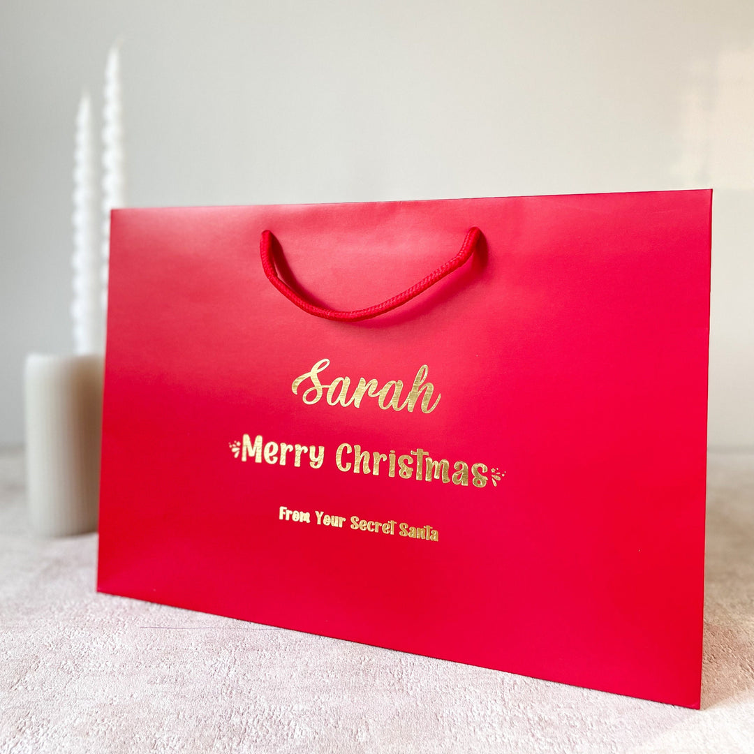 Personalized Secret Santa gift bags in red and white with custom text. The large bag measures 350x240x100mm and the medium bag measures 200x240x100mm. Both bags feature the name "Sarah" and the message "Merry Christmas" along with "From Your Secret Santa."