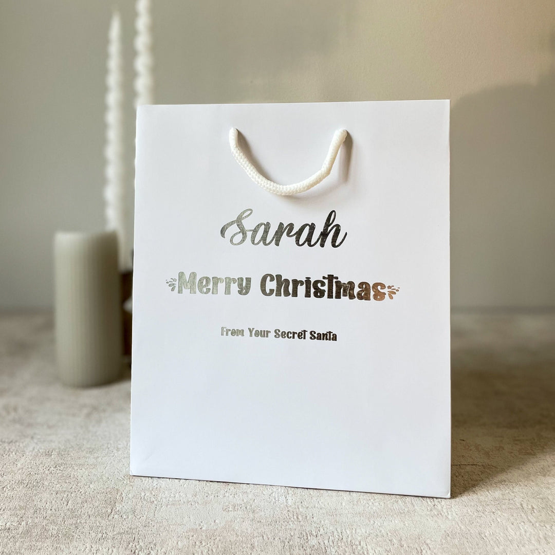A white gift bag with rope handles, featuring Secret Santa Gift Bags personalized text 'Sarah' and 'Merry Christmas' from 'Your Secret Santa'.